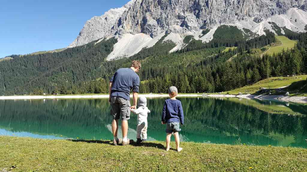 Alpenkulisse mit See, davor in Rückenansicht ein Vater mit zwei kleinen Kindern in idyllischer Landschaft. Vereinbarkeit von Familie und Beruf ist eine große Herausforderung, aber machbar. Nadja Luge von "Mama im Spagat" im Montagsinterview.